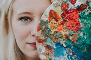 Eine Malerin schaut hinter einer bemalten Palette bunter Farben hervor